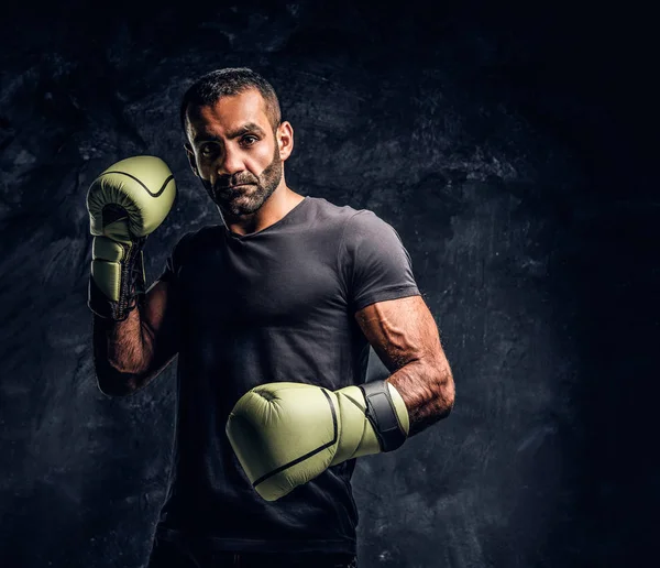 Portrait of a brutal professional fighter in a black shirt and gloves. Studio photo against a dark textured wall — Stock Photo, Image