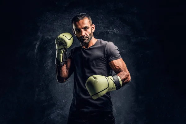 Portrait of a brutal professional fighter in a black shirt and gloves. Studio photo against a dark textured wall — Stock Photo, Image