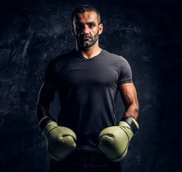 Portrait of a brutal professional fighter in a black shirt and gloves. Studio photo against a dark textured wall — Stock Photo, Image