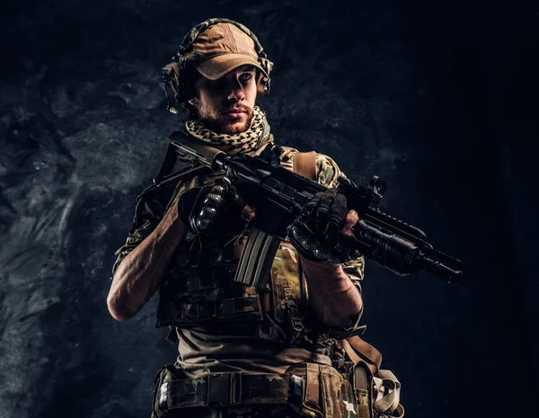 Soldado totalmente equipado con uniforme de camuflaje que sostiene un rifle de asalto. Foto del estudio contra una pared oscura — Foto de Stock