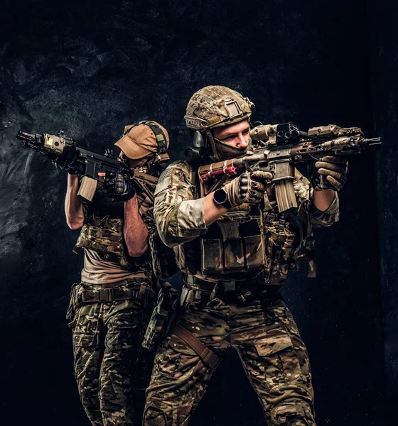 Due soldati delle forze speciali in pieno equipaggiamento protettivo con fucili d'assalto e mirano ai bersagli. Studio foto contro un muro scuro strutturato . — Foto Stock