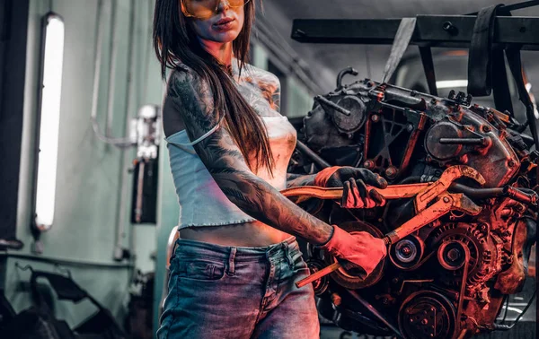 Стильная женская модель с татуированным кузовом ремонтирует автомобильный двигатель, подвешенный на гидравлическом подъемнике в мастерской. Фото с красной подсветкой — стоковое фото