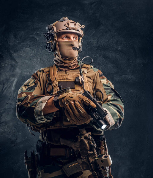 Special forces soldier in camouflage uniform holding a gun with a flashlight and looking sideways. Studio photo against a dark textured wall