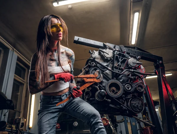 Stylowy model kobiece naprawy silnika samochodu, zawieszone na podnośniku hydraulicznym w warsztacie. — Zdjęcie stockowe