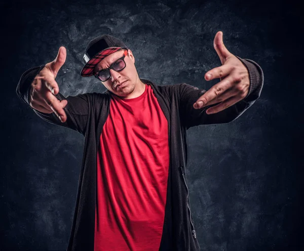 Retrato de un joven rapero vestido en un estilo hip-hop, posando para una cámara. Foto del estudio contra una pared oscura —  Fotos de Stock