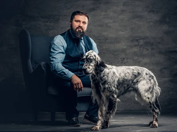 Caçador barbudo vestindo roupas elegantes sentado em um sofá com seu cão de caça branco Inglês . — Fotografia de Stock