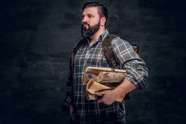 Retrato de um lenhador barbudo com uma mochila vestida com uma camisa xadrez segurando lenha . — Fotografia de Stock