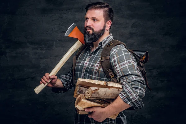一个长胡子的伐木工人的肖像, 他的背包里穿着格子衬衫, 手里拿着柴火和斧头. — 图库照片