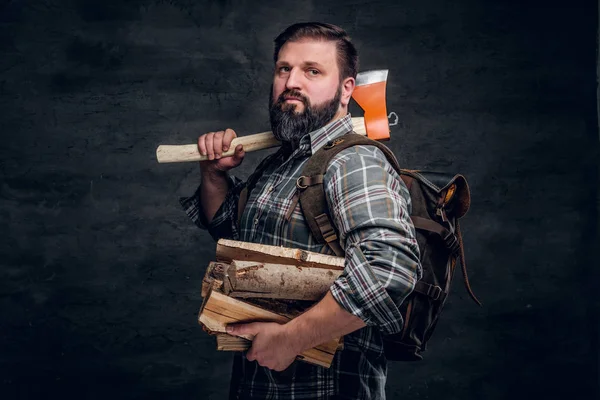 Retrato de um lenhador barbudo com uma mochila vestida com uma camisa xadrez segurando lenha e machado . — Fotografia de Stock