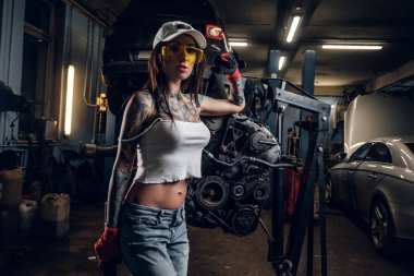 Baştan çıkarıcı tattoed kız kap ve hidrolik vinç atölye üzerinde askıya bir araba motoru yanında poz kirli giysiler giyiyor