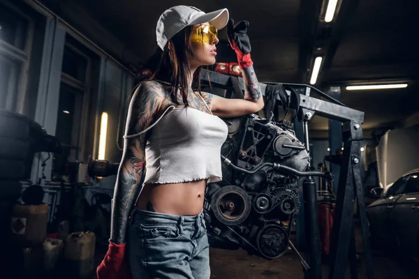 Séduisante fille tatouée portant un chapeau et des vêtements sales posant à côté d'un moteur de voiture suspendu à un palan hydraulique dans l'atelier — Photo