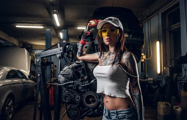 Seductive tattoed girl wearing cap and dirty clothes posing next to a car engine suspended on a hydraulic hoist in the workshop — Stock Photo, Image