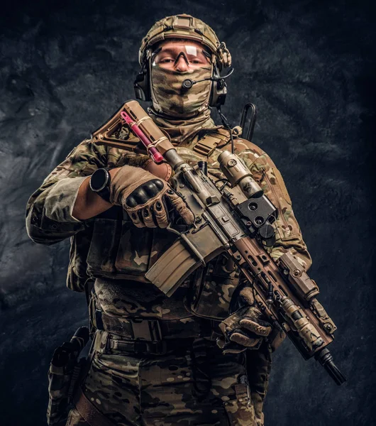 Soldado totalmente equipado con uniforme de camuflaje que sostiene un rifle de asalto. Foto del estudio contra una pared oscura — Foto de Stock