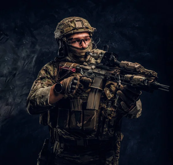 La unidad de élite, soldado de las fuerzas especiales en uniforme de camuflaje que sostiene un rifle de asalto con una mira láser y apunta al objetivo. Foto del estudio contra una pared oscura — Foto de Stock