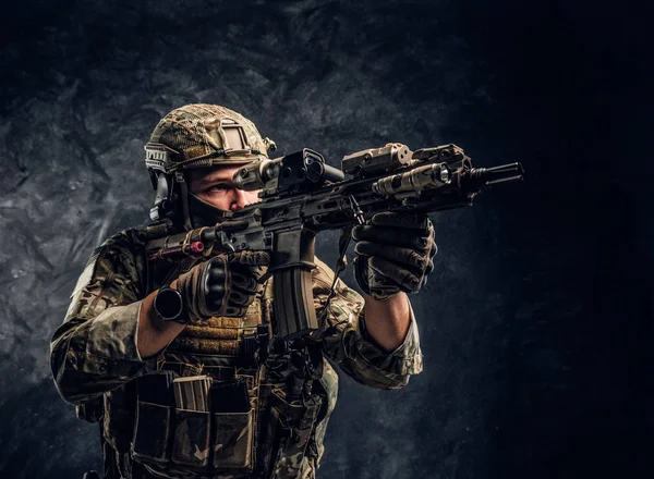 A unidade de elite, soldado das forças especiais em uniforme de camuflagem segurando um rifle de assalto com uma mira de laser e aponta para o alvo. Foto de estúdio contra uma parede escura — Fotografia de Stock