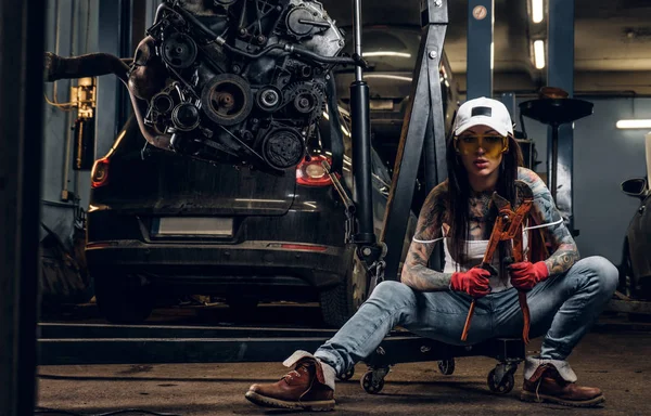 Elegante chica tatuada sosteniendo una llave grande sentada en un polipasto hidráulico con un motor de coche suspendido en el taller —  Fotos de Stock