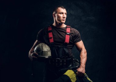 Brutal muscular fireman holding a helmet and jacket standing in the studio against a dark textured wall clipart
