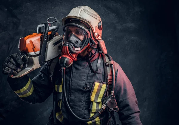 Firefighter wearing full protective equipment posing with a chainsaw on his shoulder. Studio photo against a dark textured wall — Stock Photo, Image