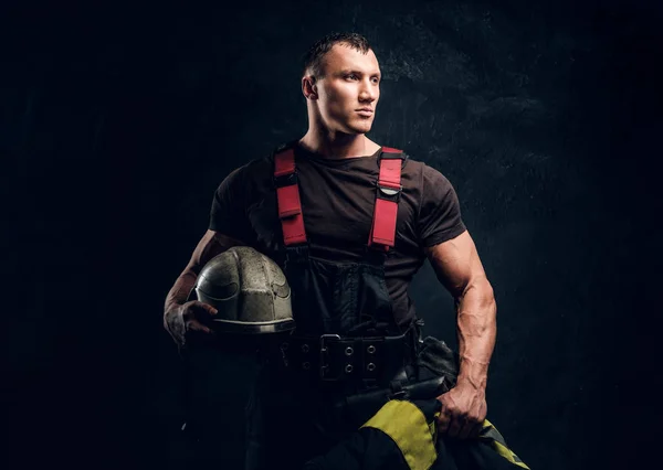 Brutal bombeiro muscular segurando um capacete e casaco de pé no estúdio contra uma parede escura texturizada — Fotografia de Stock