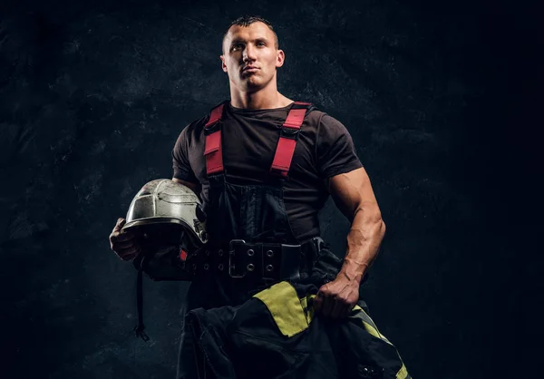 Brutal muscular fireman holding a helmet and jacket standing in the studio against a dark textured wall — Stock Photo, Image