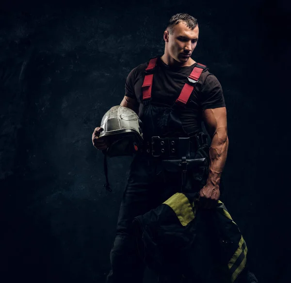 Brutal bombeiro muscular segurando um capacete e casaco de pé no estúdio contra uma parede escura texturizada — Fotografia de Stock