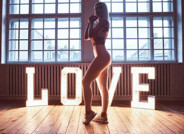 Schöne Fitness-Model trägt Sportbekleidung posiert in der Nähe von großen beleuchteten Wort Liebe in einem Raum mit Lift-Interieur. — Stockfoto