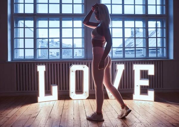 Nádherné fitness model nosí sportovní lodě poblíž velké Illuminated slovo láska v místnosti s výtahem interiér. — Stock fotografie