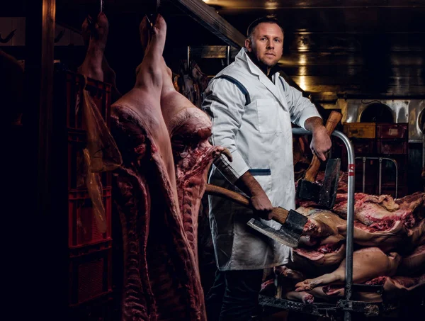 Açougueiro em vestuário de trabalho posando com dois eixos em um armazém refrigerado no meio de carcaças de carne — Fotografia de Stock