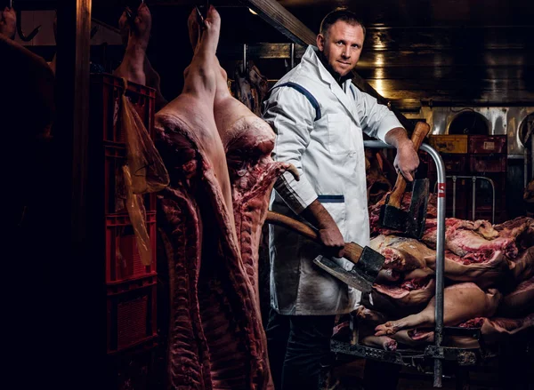 Butcher in workwear posing with two axes in a refrigerated warehouse in the midst of meat carcasses