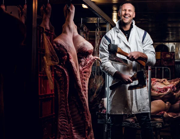 Açougueiro em vestuário de trabalho posando com dois eixos em um armazém refrigerado no meio de carcaças de carne — Fotografia de Stock