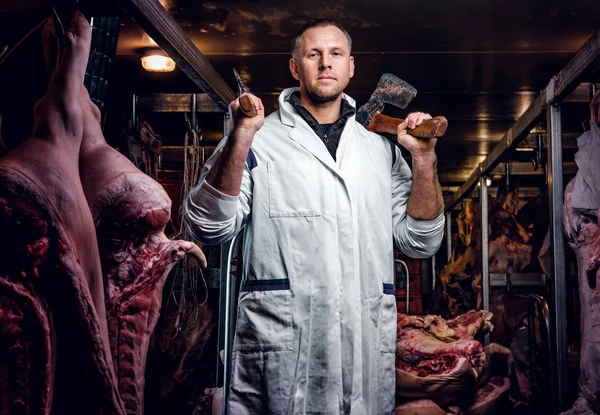 Açougueiro em vestuário de trabalho posando com dois eixos em um armazém refrigerado no meio de carcaças de carne — Fotografia de Stock