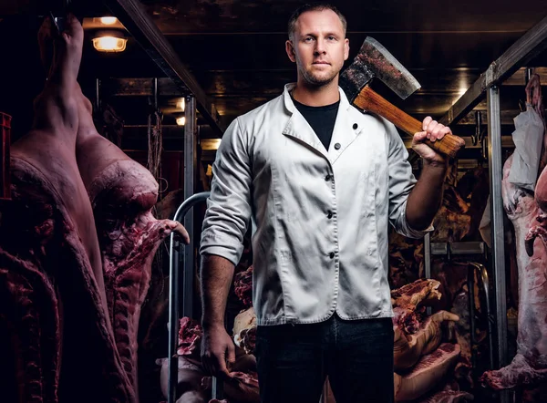 Butcher in a white work shirt holding ax while standing in a refrigerated warehouse in the midst of meat carcasses — Stock Photo, Image