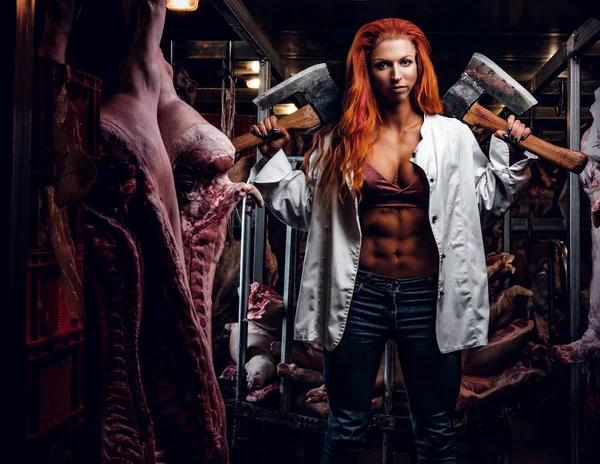 Redhead fitness donna che indossa camicia bianca sbottonata posa con due asce in un magazzino refrigerato in mezzo a carcasse di carne — Foto Stock