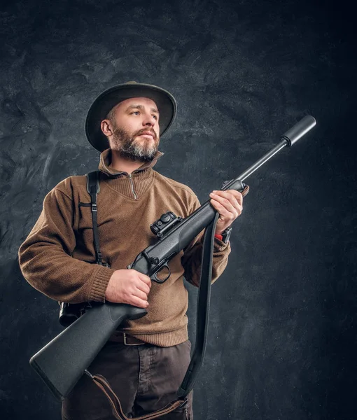 Portrait of a bearded hunter holding a rifle and looking sideways. Studio photo against dark wall background — Stock Photo, Image