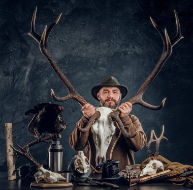Şapkalı bir büyük geyik kafatası ekipman ve trophys içeren bir tablo yanında dururken tutan olgun sakallı avcı.