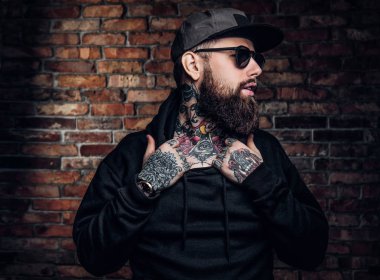 Bir siyah hoodie ve güneş gözlüğü bir şık dövmeli adam. Studio fotoğraf duvara karşı