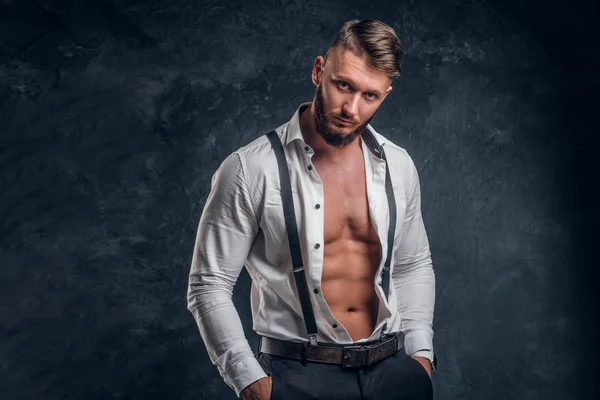 Un giovane elegante con un corpo muscoloso nella camicia bianca sbottonata con bretelle. Studio foto contro sfondo scuro parete — Foto Stock