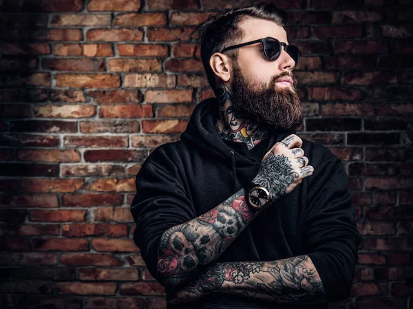 Un hombre tatuado con estilo en una sudadera con capucha negra y gafas de sol. Estudio foto contra pared de ladrillo —  Fotos de Stock