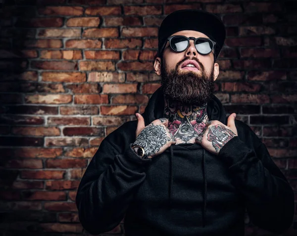 A stylish tattooed guy in a black hoodie and sunglasses. Studio photo against brick wall — Stock Photo, Image