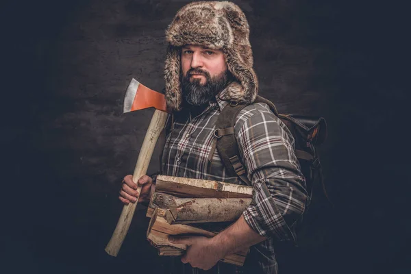 Retrato de um lenhador barbudo com uma mochila vestida com uma camisa xadrez e chapéu de caçador segurando lenha e machado . — Fotografia de Stock