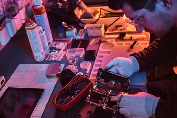 O técnico usa uma lupa para inspecionar cuidadosamente as partes internas do smartphone em uma oficina de reparo moderna. Iluminação com luzes vermelhas e azuis — Fotografia de Stock