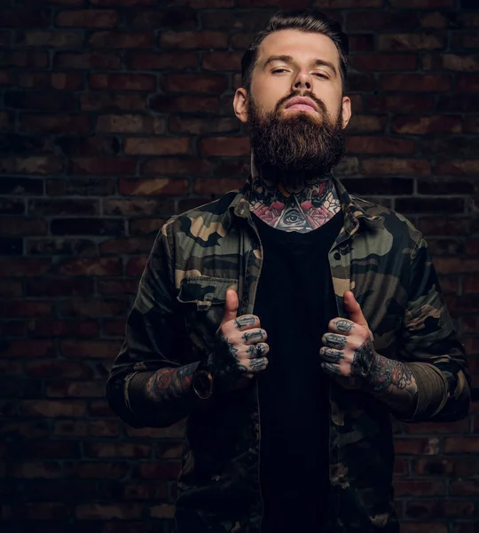 Confident bearded guy with tattoos on his hands and neck in the military shirt. Studio photo against brick wall — Stock Photo, Image