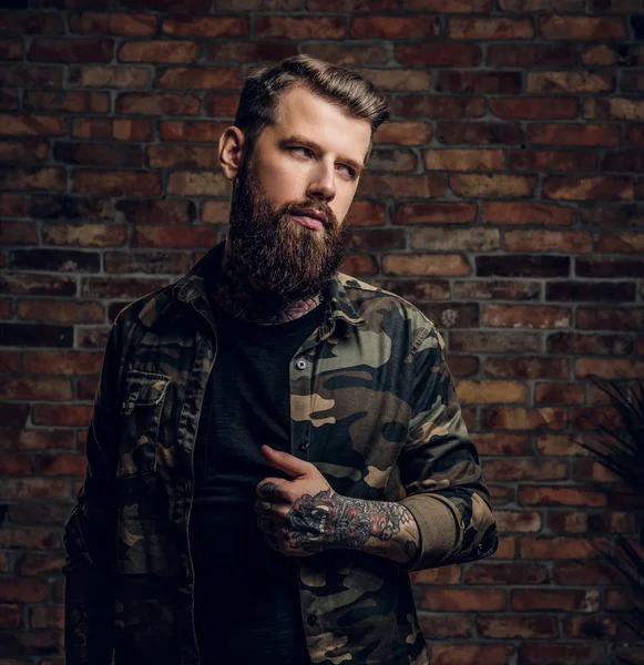Портрет скучающего татуированного бородатого парня в военной рубашке. Студийное фото на фоне стены — стоковое фото