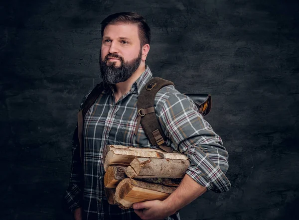 Retrato de um lenhador barbudo com uma mochila vestida com uma camisa xadrez segurando lenha . — Fotografia de Stock