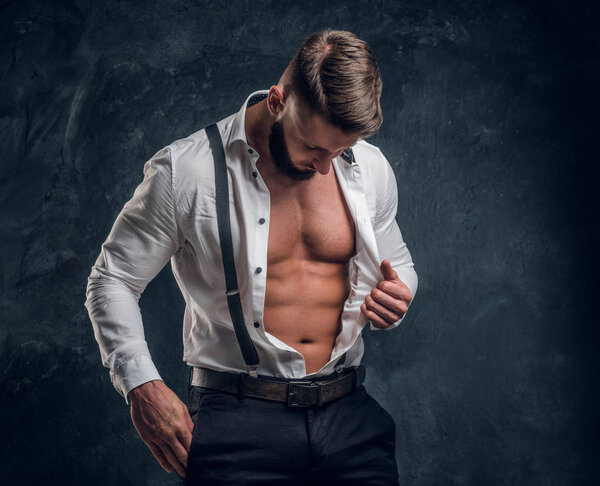 Стильный молодой человек с мускулистым телом в расстегнутой белой рубашке с подтяжками. Студийное фото на фоне темной стены
