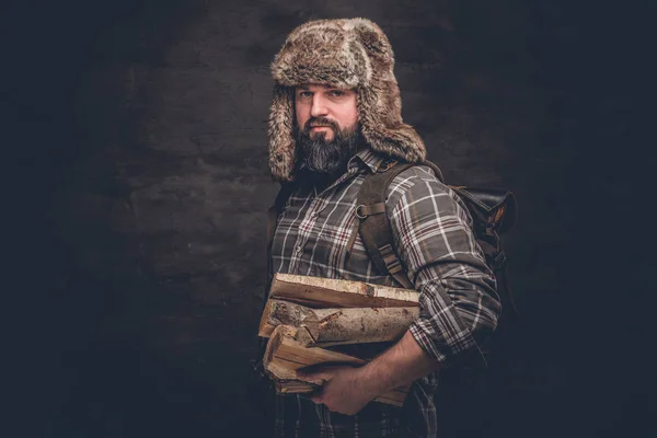 Retrato de um lenhador barbudo com uma mochila vestida com uma camisa xadrez e chapéu de caçador segurando lenha . — Fotografia de Stock