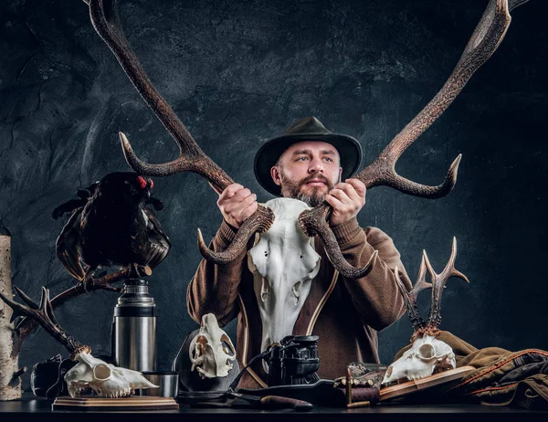 Érett szakállas vadász gazdaság egy nagy szarvas koponya mellett egy asztal eszközök és trophys állva kalapban. — Stock Fotó