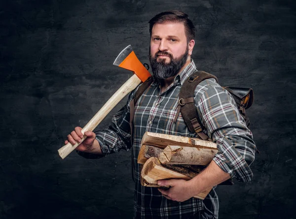 Porträt eines bärtigen Holzfällers mit Rucksack in einem karierten Hemd mit Brennholz und Axt. — Stockfoto