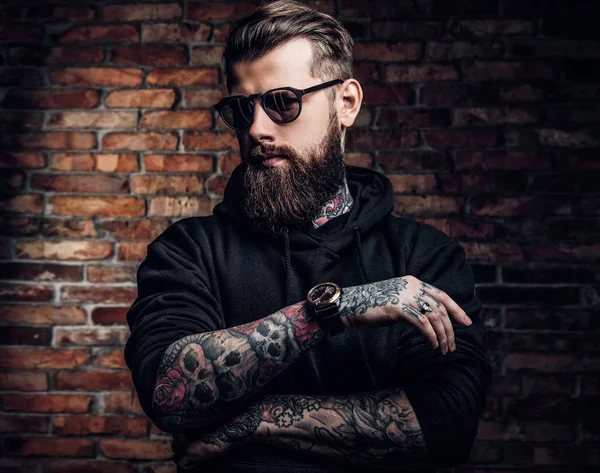 Un mec tatoué élégant avec un sweat à capuche noir et des lunettes de soleil. Studio photo contre mur de briques — Photo