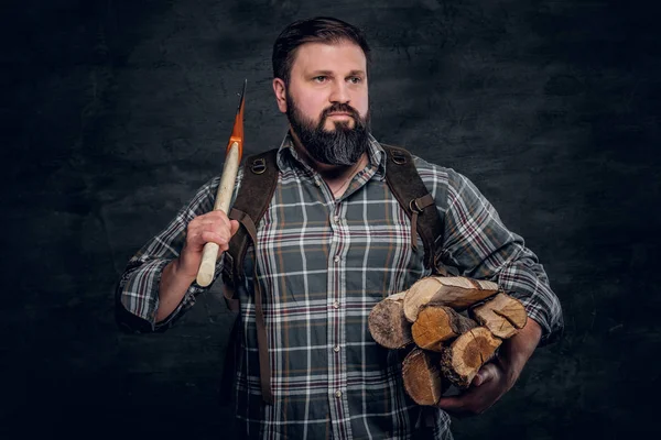 Portrait of a bearded woodcutter with a backpack dressed in a plaid shirt holding firewood and ax. — Stock Photo, Image